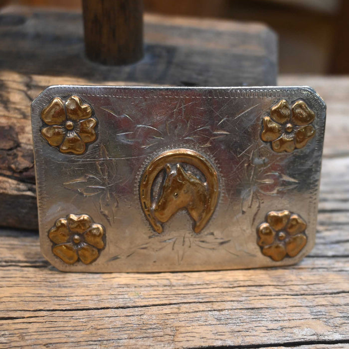 Metal belt buckle with floral designs and a horse head on a wooden surface